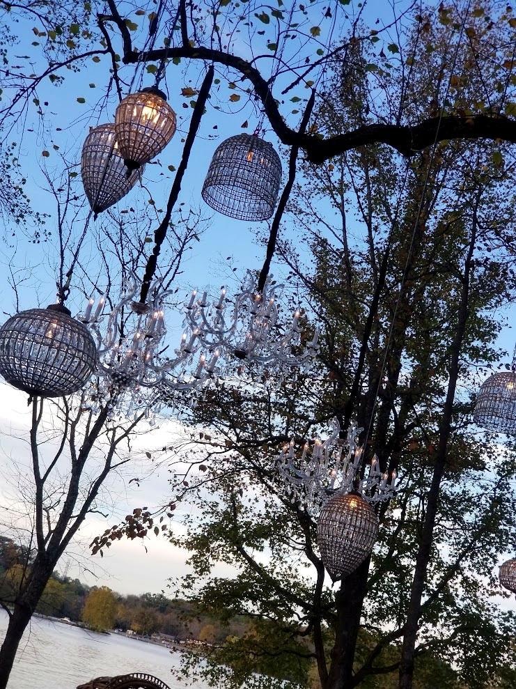 Outdoor chandeliers and globe lanterns hanging in trees at waterfront dusk